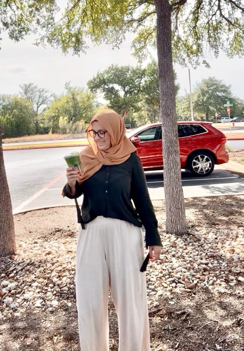 A woman holding a green juice, standing under trees on a bright afternoon