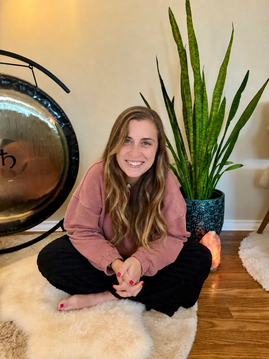 Kelsey Craft sitting cross-legged on a sheepskin beside a gong and a snake plant