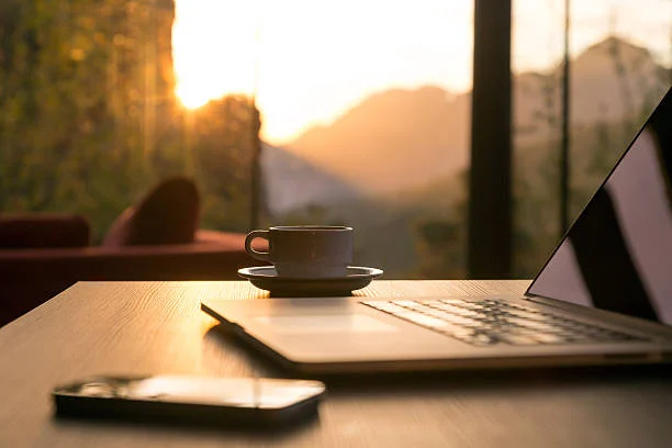 A laptop and a cup of coffee on a desk, sunrise over mountains through the window